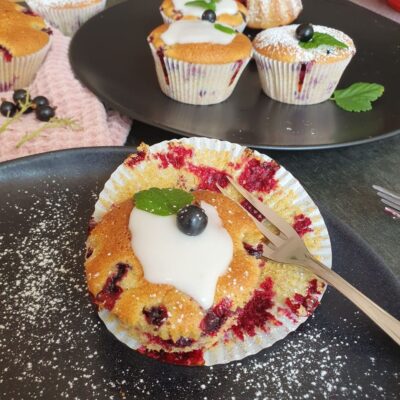 Einkorn Cassis Muffin mit Zuckerglasur und Dekobeeren und Blatt. mit Kuchengabel auf schwarzem Teller mit etwas Puderzucker