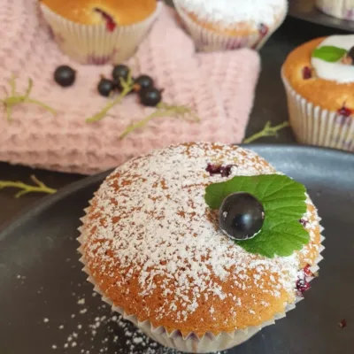 Einkorn Cassis Muffin mit Puderzucker bestäubt. Johannisbeere und Blatt als Deko. Dahinter ein rosa Tuch und weitere Beeren