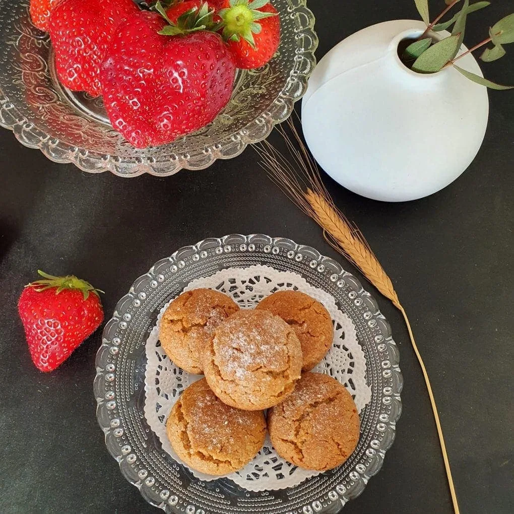 fünf Kekse auf Glasteller auf weißem Papier auf Glasteller. Ähre daneben. Glasschale mit Erdbeeren und weiße Vase daneben auf schwarzem Grund