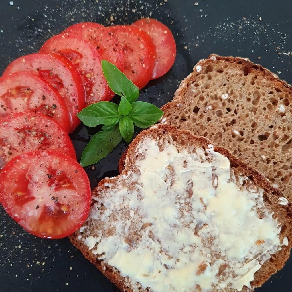 Urkornbrot mit Butter bestrichen, Tomaten und Basilikum liegen daneben auf schwarzem Untergrund