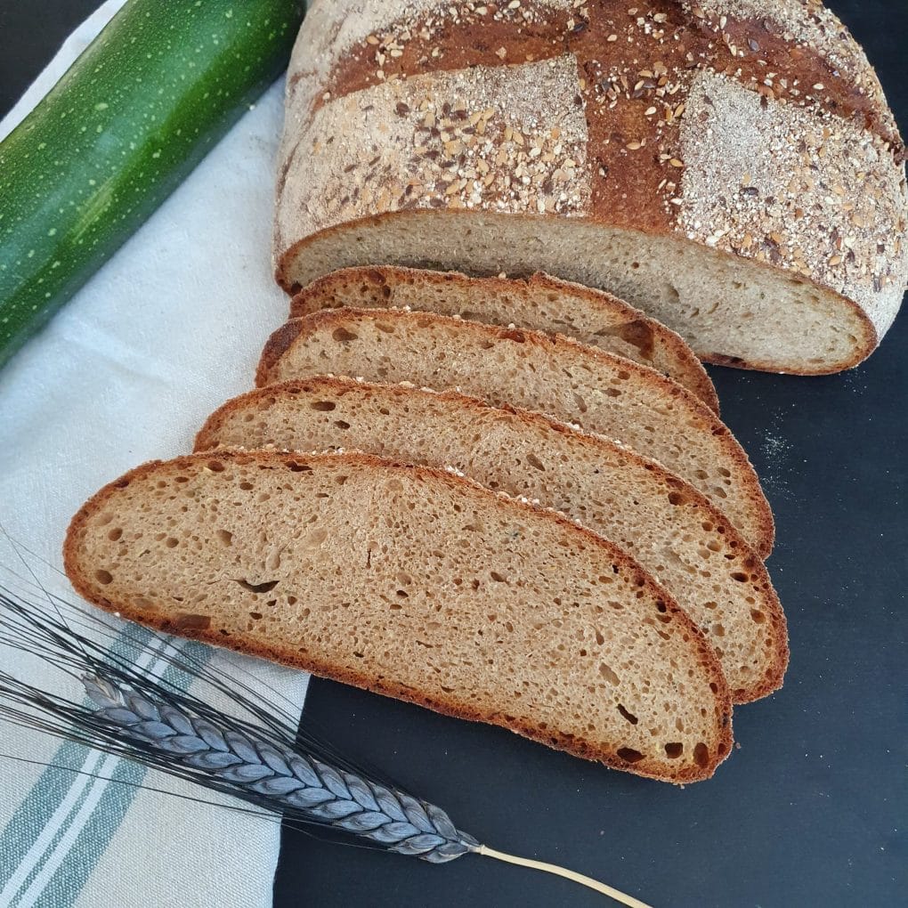 vier Brotscheiben liegen vor einem angeschnittenen Laib Brot. Im Hintergrund eine grüne Zucchini