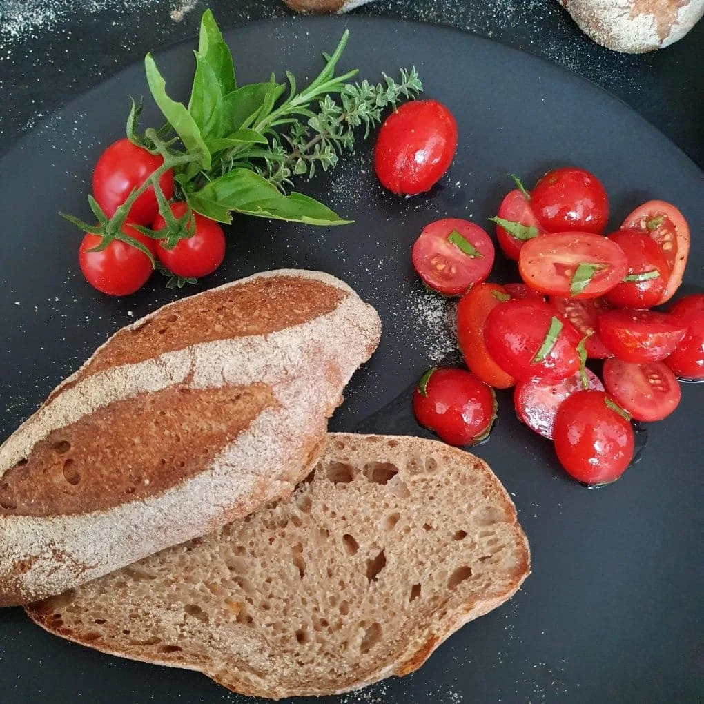 Urkornbrötchen aNette liegt aufgeschnitten auf einem schwarzen Teller. Daneben Kirschtomatensalat und Gartenkräuter