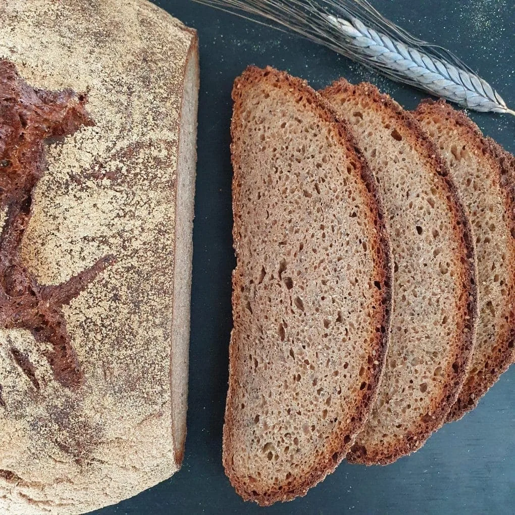Ausschnitt eines runden Brotlaibes von oben. Daneben 3 Brotscheiben und eine Ähre