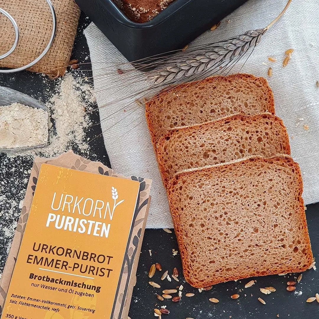 EmmerPurist fertig gebacken und aufgeschnitten auf dem Tisch. Daneben eine aufgerissene Packung, Getreideähre und Deko