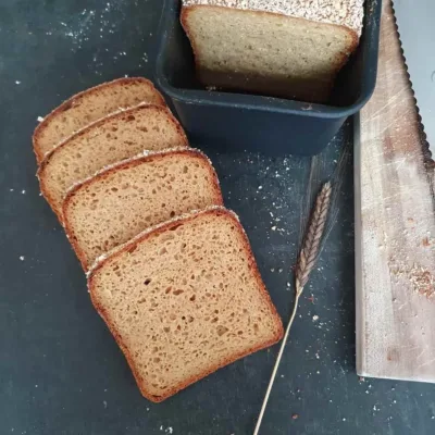 4 Brotscheiben Einkorn Vollkornbrot mit Buttermilch und Kartoffeln. Daneben eine schwarze Brotform mit dem angeschnittenen Brotlaib
