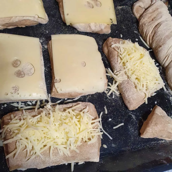 Teigstücke für Brötchen mit Käsebestreut liegen auf einem schwarzen Backblech