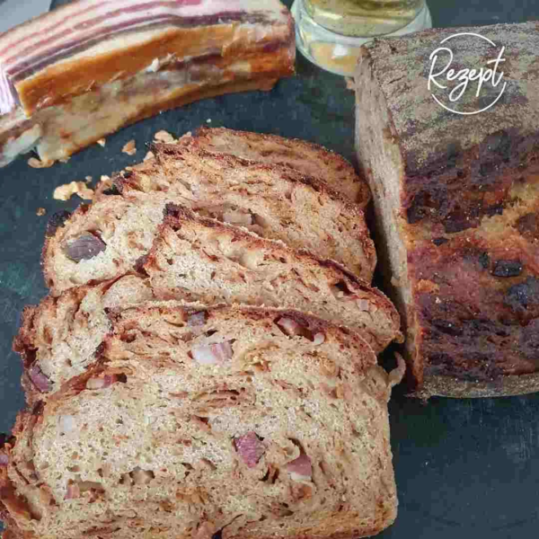 Urkorn Männerbrot mit Speck und Zwiebel. Viel Scheiben aufgeschnitten neben Brotlaib. Daneben ein Stück Speck