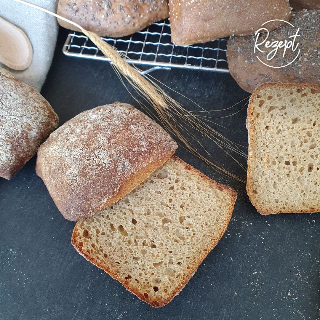 einkornbrötchen version III aufgeschnitten mit Schnittseite nach oben auf schwarzem Tisch. Daneben Einkornähre