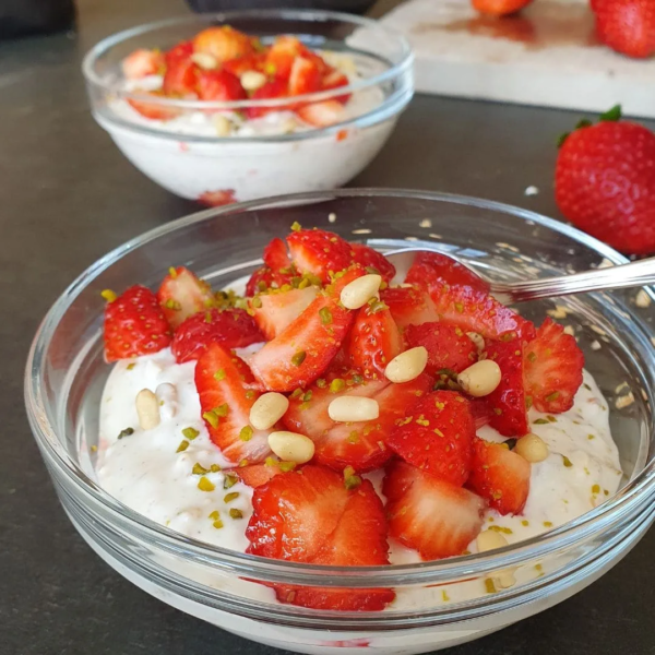 Erdbeer-Urkorn-Overnight in einer Glasschale und Löffel fertig zum Servieren auf schwarzem Tisch. Im hintergrund eine weitere gefüllte Frühstücksschale