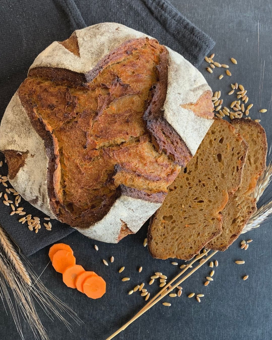 Karotten Sauerteigbrot liegt angeschnitten auf schwarzem Tisch. daneben Getreideähren, Körner, Karottenscheiben