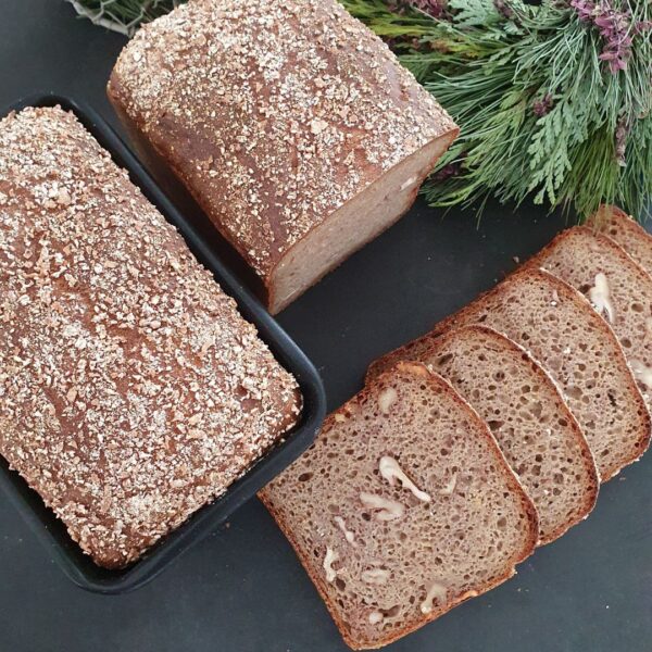 Einkornbrot mit Walnüssen in Scheiben geschnitten auf schwarzem Untergrund. Deko aus Tannenreisig daneben. Brotlaib von oben in schwarzer Backform