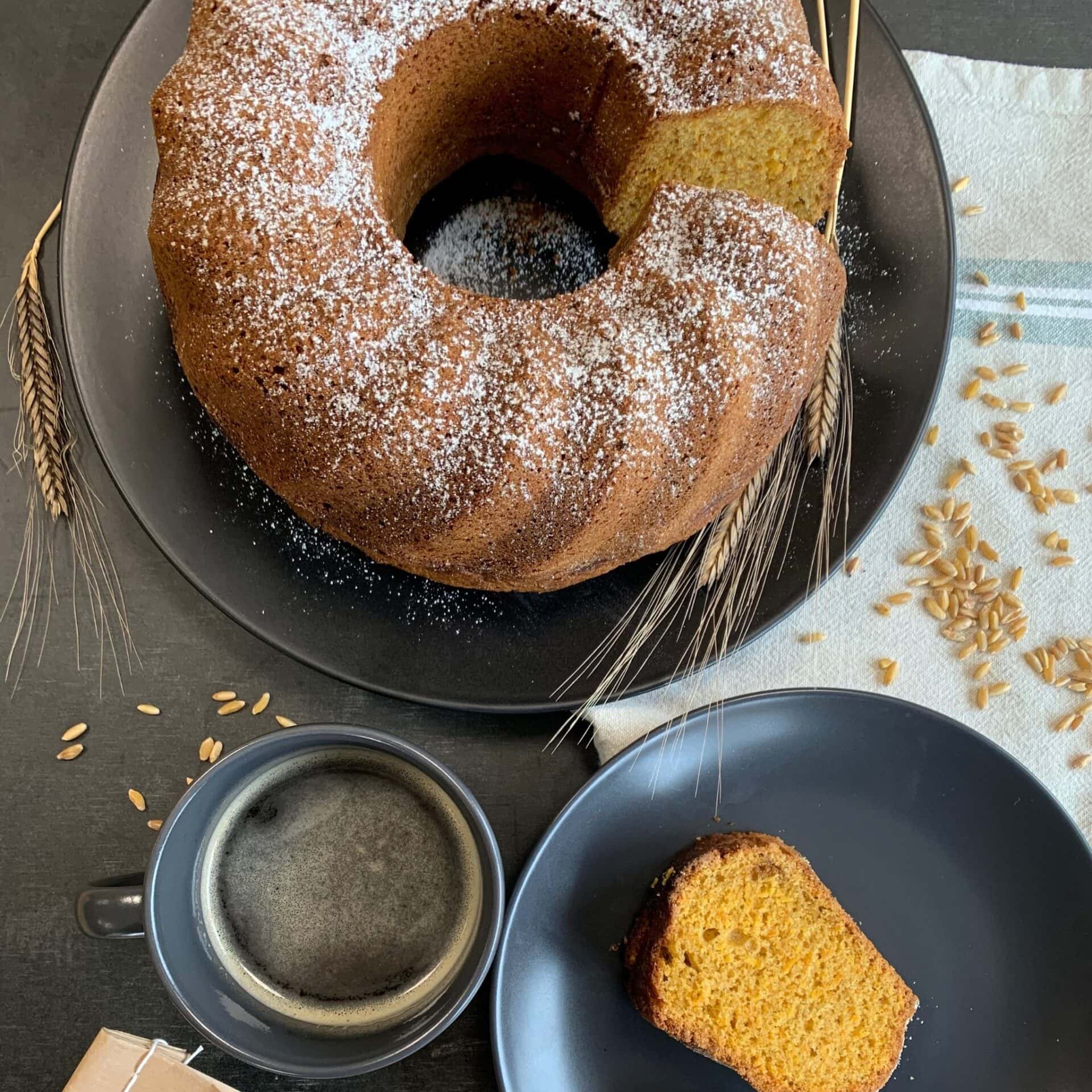 Karotten Gugelhupf auf schwarzem Teller. Körner, Kaffeetasse und graues Tuch daneben