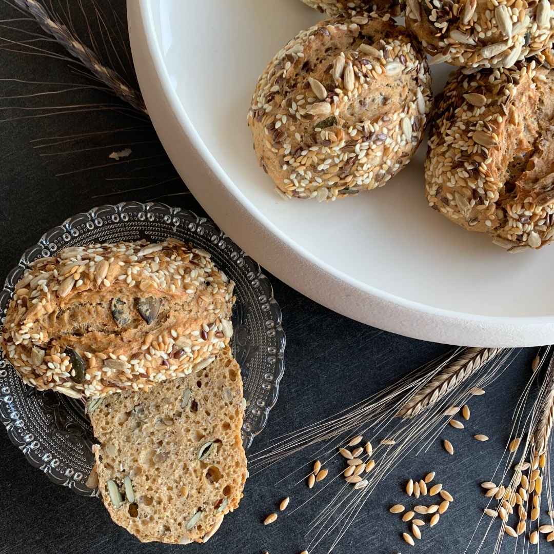 Urkornbrötchen mit Backpulver auf weißem Teller. aufgeschnittenes Brötchen auf Glasteller daneben. getreidekörner und Ähren liegen auf schwarzem Tisch