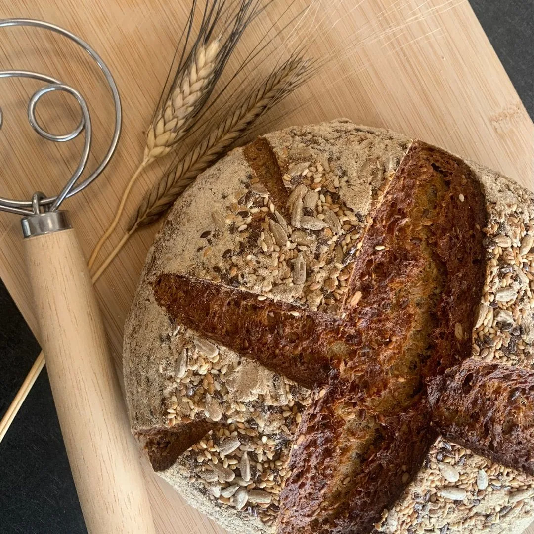 gesundes Urkornbrot aus der Brotbackmischung Buttermilchkruste. Brotlaib von oben auf einem Holzbrett. Urkornähren und ein dänischer Schneebesen daneben
