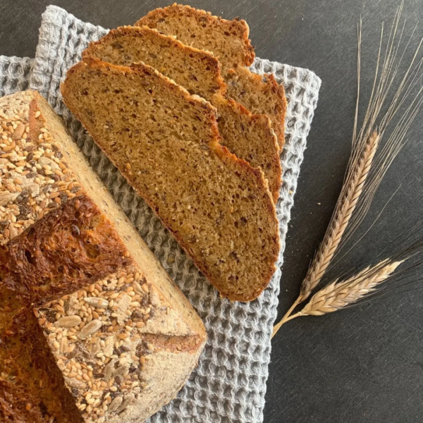 aufgeschnittener Urkorn Brotlaib von oben auf einem grauen Tuch. Brotscheiben und Urkornähren daneben auf einem schwarzen Brett