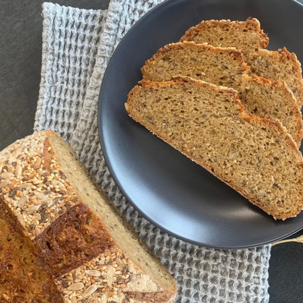 gesundes Vollkorn Saatenbrot aus der Brotbackmischung Buttermilchkruste. aufgeschnittener Brotlaib von oben auf einem grauen Tuch. Brotscheiben daneben auf einem schwarzen Teller
