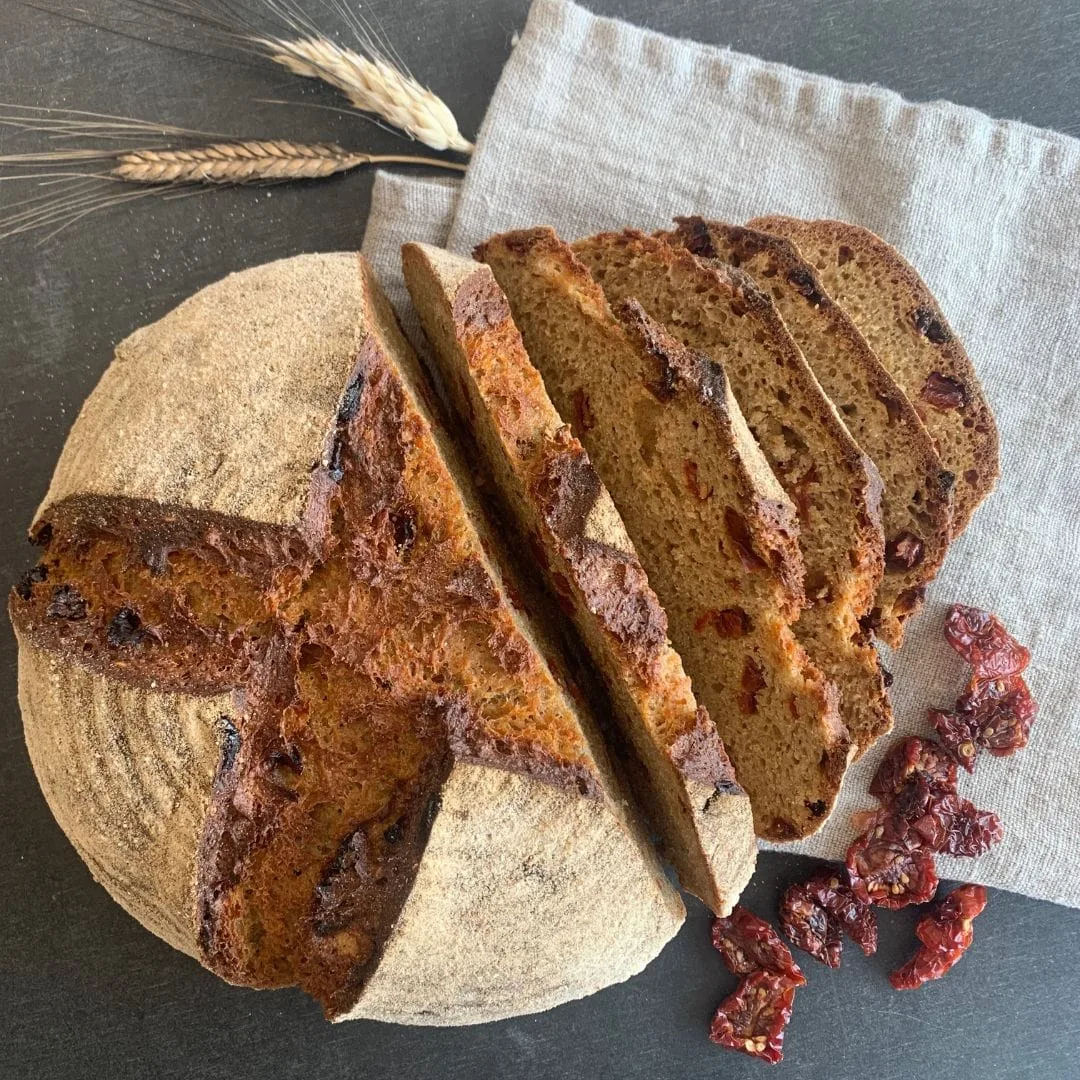 gesundes Vollkornbrot Tomatenliebe im Urkornlaib aufgeschnitten auf grauem Tuch. Daneben Ähren und getrocknete Tomaten auf schwarzem Brett