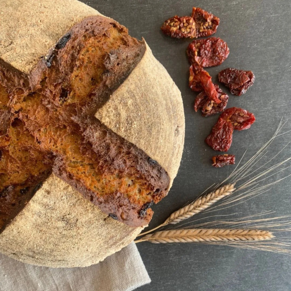 ein Laib der Tomatenliebe aus Urkornlaib ist von oben zu sehen. daneben sind zwei einkornähren und ein paar getrocknete Tomaten auf einem schwarzen Tisch zu sehen