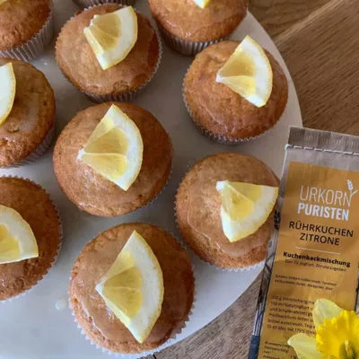 Zitronenmuffins von oben mit Zitronenscheibe garniert. Daneben liegt die Tüte einer Backmischung