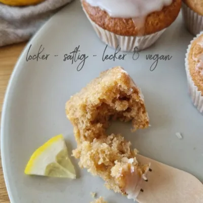 ein kleines Stück eines veganen Zitronenmuffins auf einem Teller, das mit einer Holzgabel abgetrennt wurde. Daneben liegt ein kleines Stück einer zitronenscheibe. weitere Muffins stehen am Rand des Tellers und sind ein Stück zu sehen