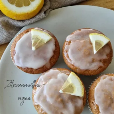 vegane Zitronenmuffins mit Zuckerguss und einem Stück Zitrone garniert stehen auf einem hellen Teller. Daneben ein Stück angeschnittene Zitrone
