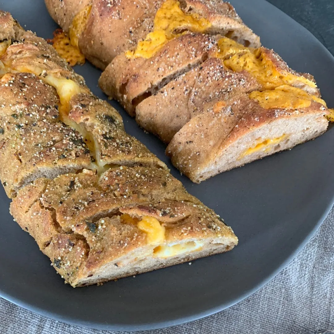aufgeschnittene Brotscheibe von BlueVelvet Käsestangen auf schwarzem Teller. Dinkelstangen sind von oben sichtbar. die Kruste leicht aufgebrochen. geschmolzener Käse sichtbar