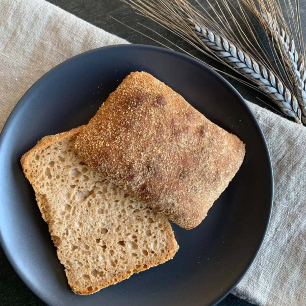 Das Brötchen Luftkornling aufgeschnitten auf schwarzem Teller. Das Teller steht auf einem beigen Tuch. Daneben liegen zwei Ähren