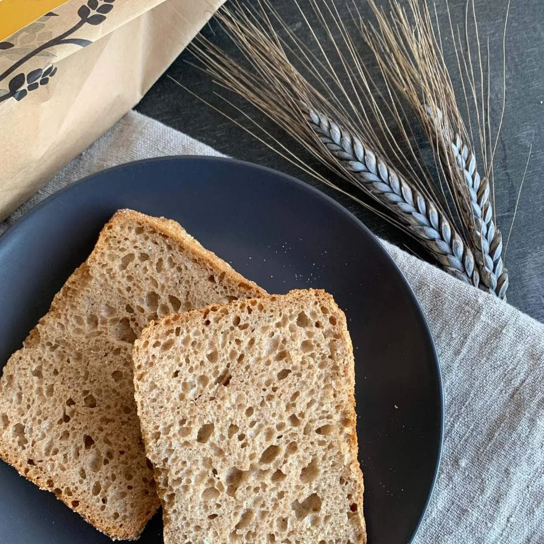 aufgeschnittenes Brötchen. Schnittfläche nach oben liegt auf schwarzem Teller. zwei Ähren und beiges Tuch daneben. In der ecke ist ein Stück einer Mehlpackung zu sehen