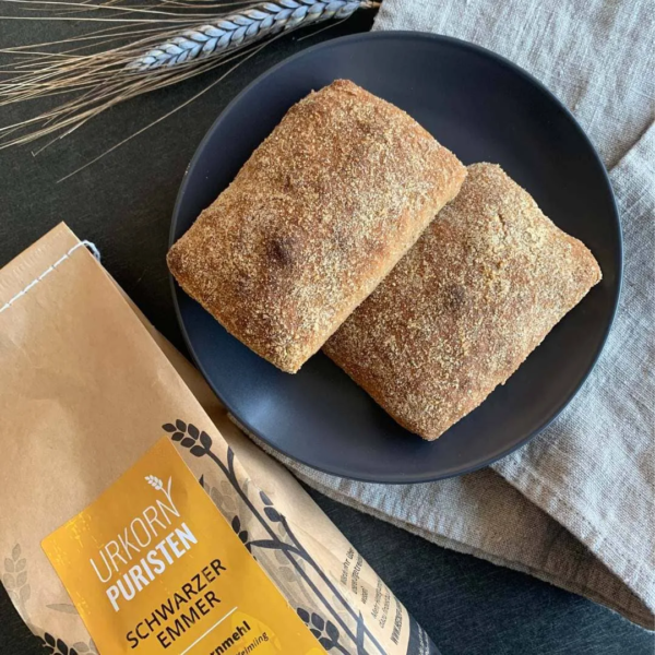 zwie stück Luftkornlinge liegen auf einem schwarzen Teller. eine Mehlpackung Emmer Vollkornmehl liegt daneben