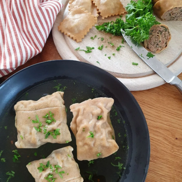 3 Urkorn Maultaschen in der Brühe liegen in einem schwarzen Suppenteller. daneben ein Holzbrett mit Messer, etwas geschnittene Petersilie, ein gestreiftes Tuch. Am Rand sind noch kleine Maultaschen zu sehen