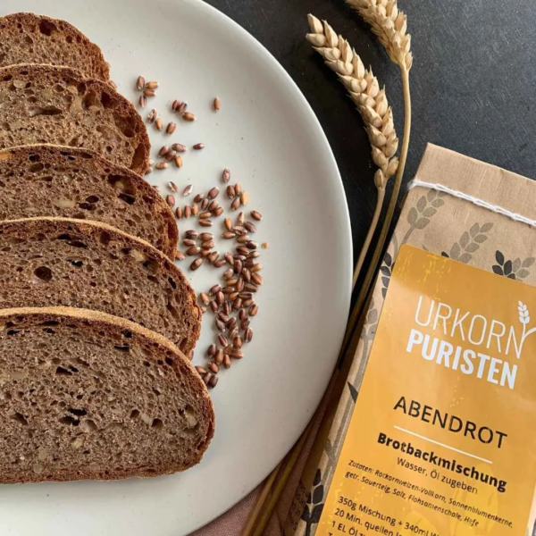 fünf scheiben Brot aus der Backmischung Abendrot auf einem beigen Teller. Daneben Getreidekörner vom Rotkornweizen. Zwei Ähren liegen auf einem schwarzen Tisch auf dem das beige Teller steht. rechts unten im Bild ein Ausschnitt einer Packung der Backmischung zu sehen