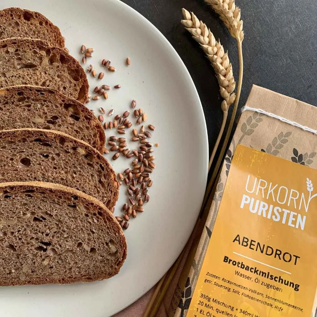 fünf scheiben Brot aus der Backmischung Abendrot auf einem beigen Teller. Daneben Getreidekörner vom Rotkornweizen. Zwei Ähren liegen auf einem schwarzen Tisch auf dem das beige Teller steht. rechts unten im Bild ein Ausschnitt einer Packung der Backmischung zu sehen