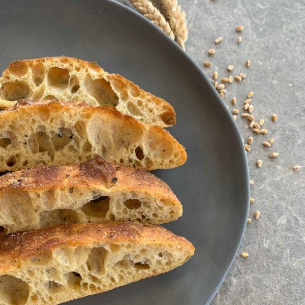 vier Scheiben Fladenbrot mit Gelbmehlweizen liegen auf einem schwarzen Teller. daneben liegen ein paar Getreidekörner und am Rand ist ein Stück einer Ähre zu erkennen