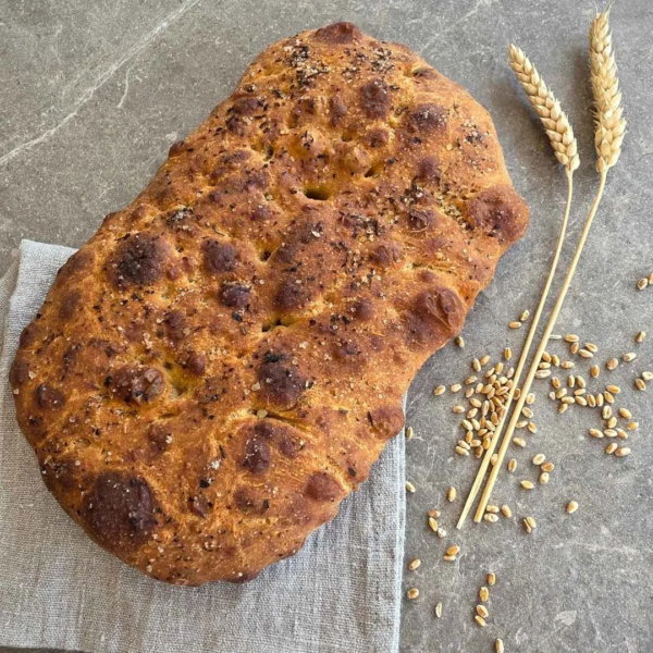 ein fertig gebackenes Fladenbrot liegt auf einem grauen Tuch. Daneben liegen 2 Getreideähren und ein paar Getreidekörner