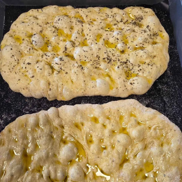 zwei Teigstücke für Fladenbrot liegt auf einem schwarzen Blech. Die Teiglinge sind mit Olivenöl bestrichen und mit Kräutersalz bestreut