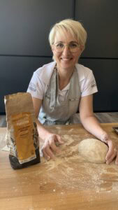 Stefanie Dehn beim Backen. Sie steht hinter einem mit Mehl bestreutem Holztisch. auf dem Tisch steht eine Mehlpackung. Ihre Hand liegt auf einer Teigkugel