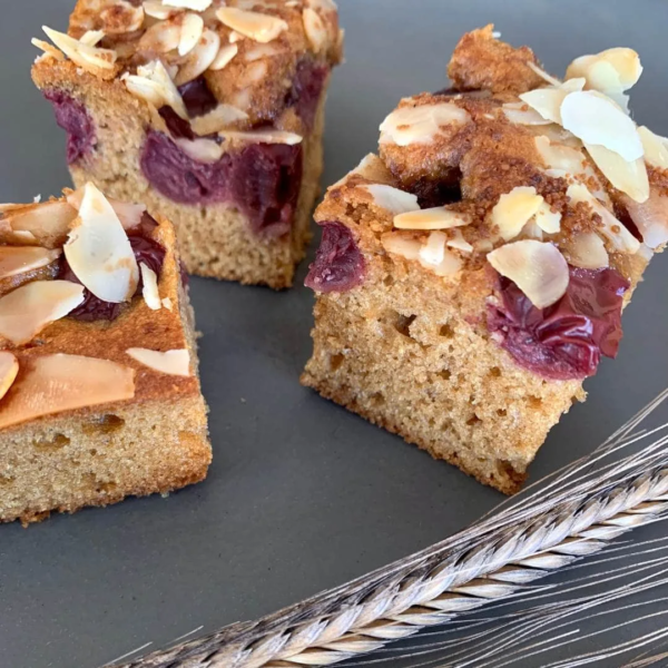 drei Stücke Kirsch-Mandelkuchen auf einem schwarzen Teller. Daneben eine Einkornähre
