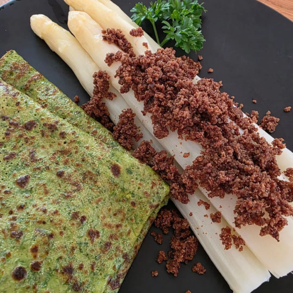 Spargel mit urkornbrotbrösel und zwei Kräuterflädle auf schwarzem Teller angerichtet. Daneben ein Stück Petersilie