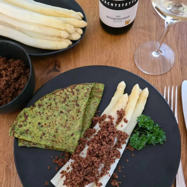 Spargel mit urkornbrotbrösel und Kräuterflädle auf schwarzem teller angerichtet. Daneben Stangespargel auf einer schwarzen Platte. Daneben ist ein stück von einem Weinglas und eine Flasche Weißwein zu sehen