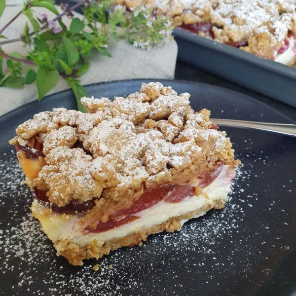 ein Stück zwetschgen Urkorn Käsekuchen auf einem schwarzen Teller mit Puderzucker bestäubt. Dahiner ein Wiesen-sommerblumenstrauß zu sehen