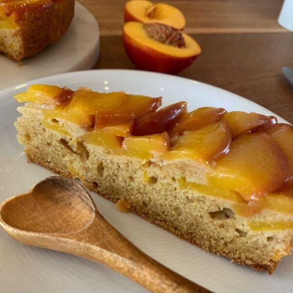 ein Kuchenstück des Aprikosenkuchen upside-down auf einem weißem Teller. Daneben liegt ein Holzlöffel in Herzform. Im Hintergund ist eine aufgeschnittene Aprikose mit Stein zu sehen und ein kleiner Ausschnitt des Kuchens der auf einer Kuchenplatte steht