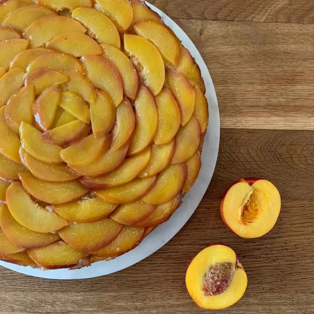Aprikosenkuchen upsida-down von oben auf einem Holztisch. Aprikosen liegen in Scheiben geschnitten fächerartig auf einem Kuchenboden. daneben zwei Hälften einer Aprikosen.