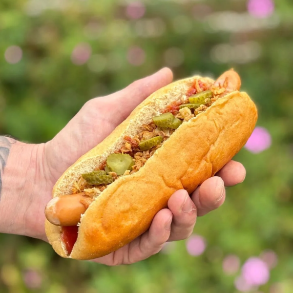 fertig angerichtetes Hotdog Brötchen in einer Hand. Im Hintergrund ist verschwommen ein Blumengarten zu sehen