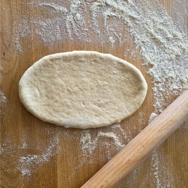 ausgerollter Teig für ein Hotdog Brötchen liegt auf einem bemehlten Holztisch. daneben ein Ausschnitt eines Rollholzes