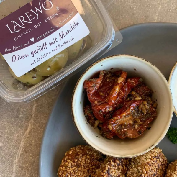 getrocknete Tomaten in einer hellen kleinen Schale. die schale steht auf einem grauen Teller. Daneben ist ein Ausschnitt von vier Stück vegane Einkorn-Falafel zu sehen. Neben dem Teller steht eine transparente dose mit eingelegten, grünen Oliven. Der Deckel der Dose hat ein lila-weißes Etikett mit der Aufschrift LAREWO