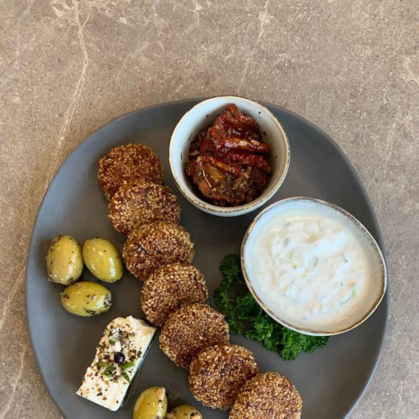 vegane Einkorn-Falafel auf einem grauen Teller angerichtet. auf dem Teller sind grüne Oliven, ein Stück Schafskäse, eine Schale mit Zsatsziki und eine schale mit getrockneten Tomaten zu sehen. Das Teller steht auf einem beigen Steintisch. der Blick ist von oben.