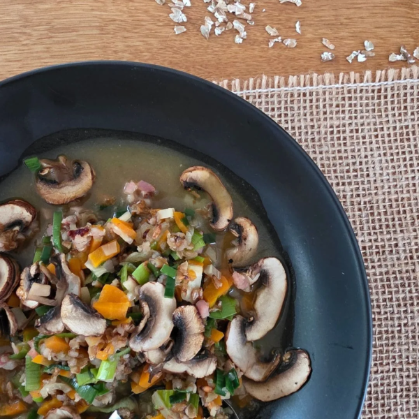 Ausschnitt von oben einer wärmende Urkorn-Gemüsesuppe in einem schwarzen Teller. Das Teller steht auf einem Juteband auf einem Holztisch. oben im Bild liegen ein paar Getreidflocken vom Waldstaudenroggen