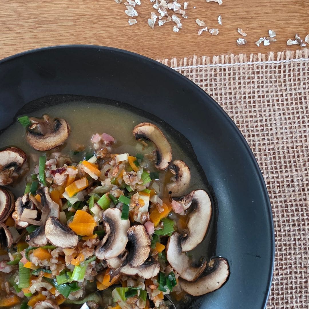 Ausschnitt von oben einer wärmende Urkorn-Gemüsesuppe in einem schwarzen Teller. Das Teller steht auf einem Juteband auf einem Holztisch. oben im Bild liegen ein paar Getreidflocken vom Waldstaudenroggen