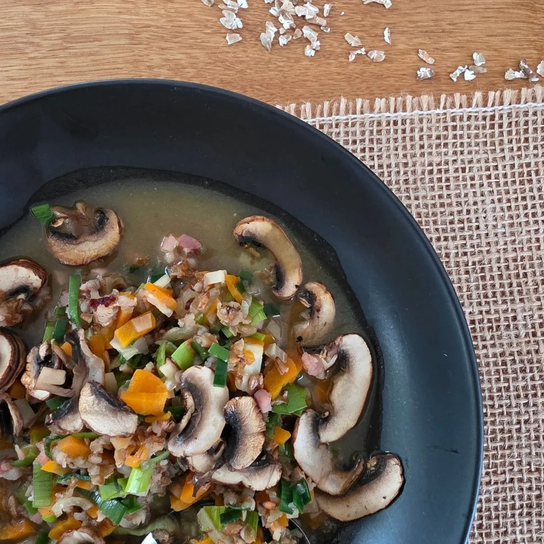 Ausschnitt von oben einer wärmende Urkorn-Gemüsesuppe in einem schwarzen Teller. Das Teller steht auf einem Juteband auf einem Holztisch. oben im Bild liegen ein paar Getreidflocken vom Waldstaudenroggen