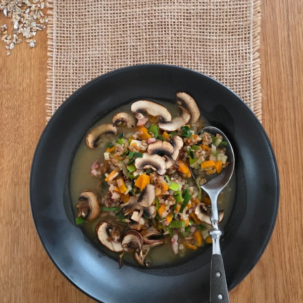 herzhaft-wärmende Urkorn-Gemüsesuppe in schwarzem Teller. Das Teller steht auf einem Holztisch auf einem Juteband. in der oberen linken Ecke sind Getreideflocken zu sehen